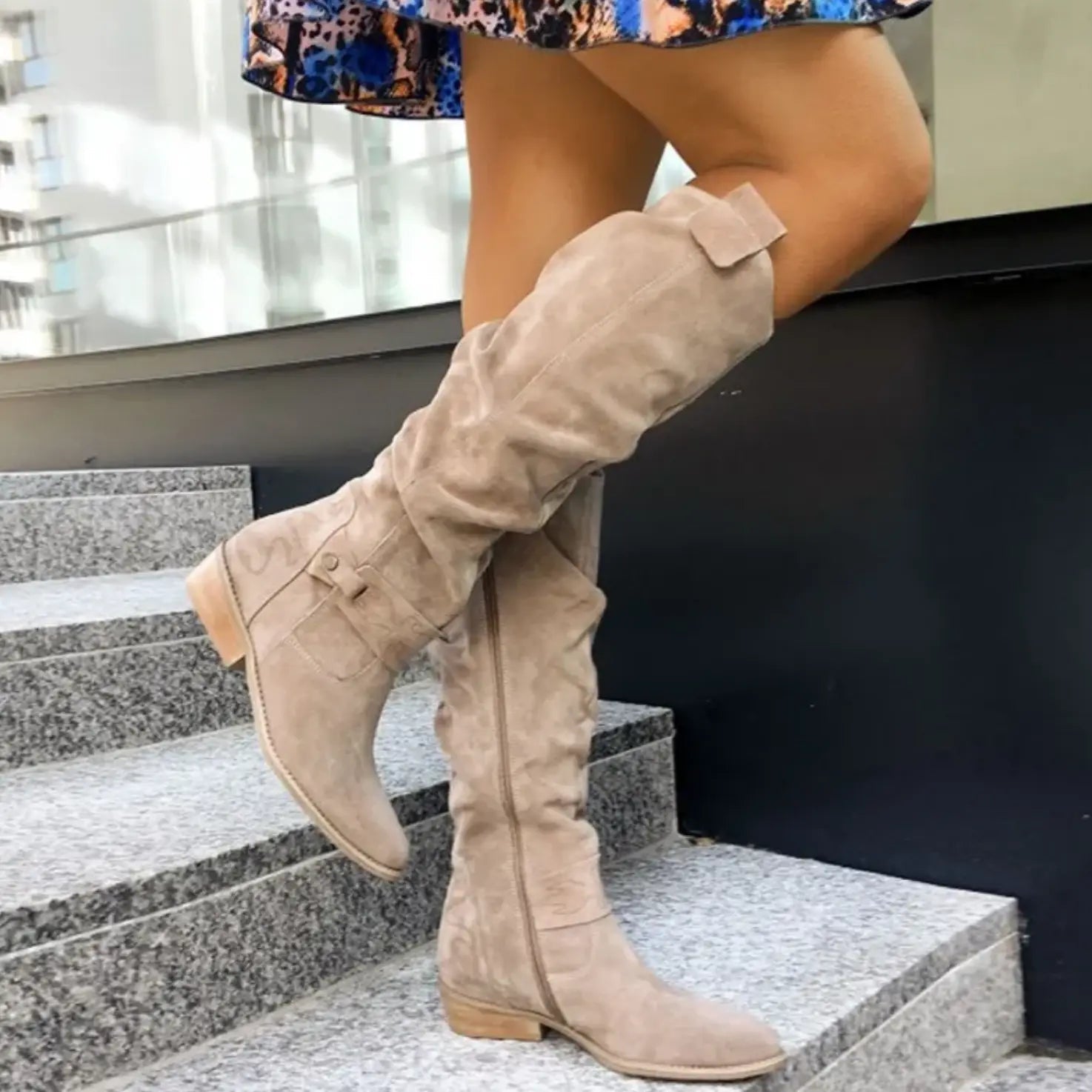 Stivali alti in camoscio beige da donna su gradini di pietra, moda autunno, tacco basso, abbinati a gonna colorata.