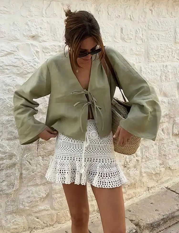 Donna con top verde oliva e gonna bianca all'uncinetto, borsa di paglia, occhiali da sole, sfondo di muro in pietra. Moda estiva casual chic.