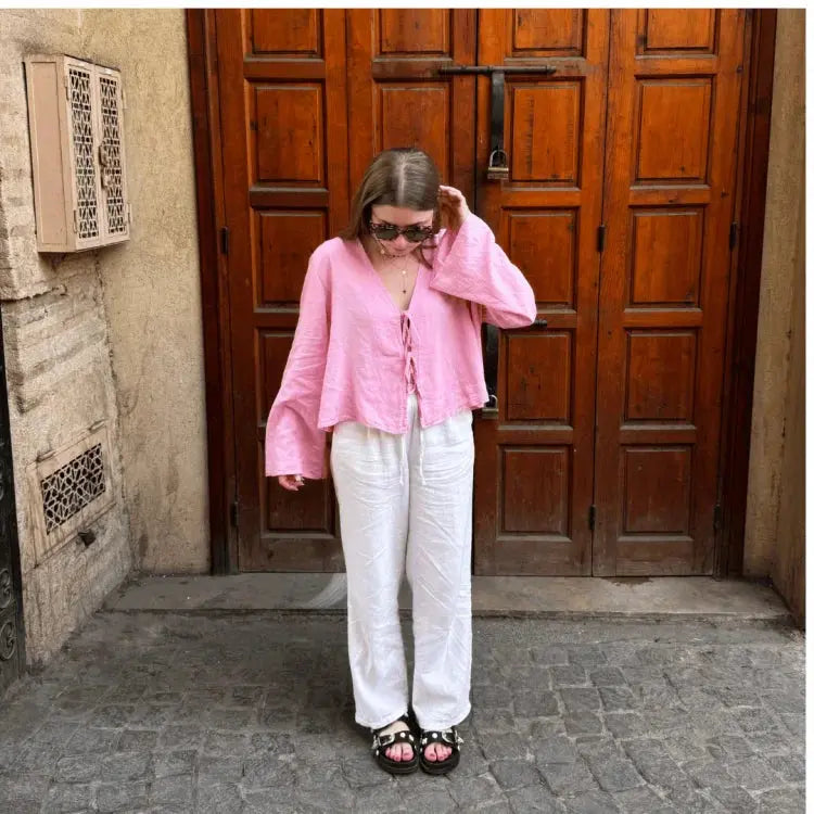 Donna con camicia rosa e pantaloni bianchi davanti a porta in legno. Indossa occhiali da sole e sandali neri. Moda estiva casual, street style.