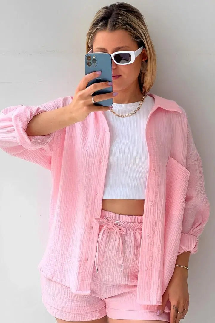 Donna con camicia rosa oversize, top bianco e shorts coordinati, indossa occhiali da sole bianchi e tiene uno smartphone. Moda estiva casual.