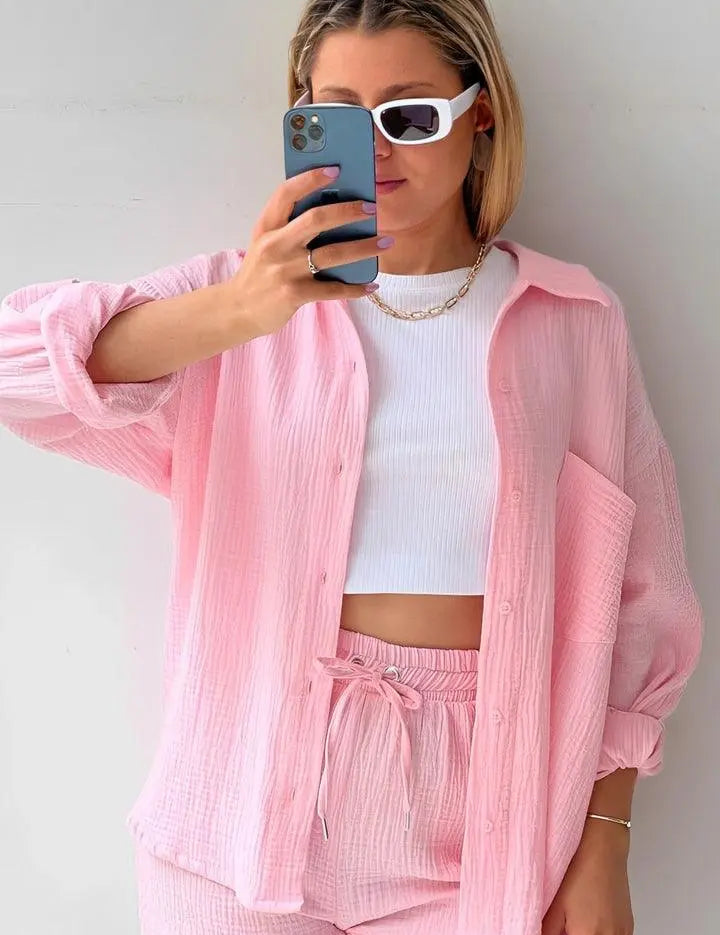 Donna con camicia rosa oversize, top bianco e shorts coordinati, indossa occhiali da sole bianchi e tiene uno smartphone. Moda estiva casual.