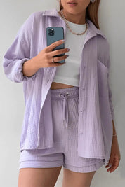 Donna con completo coordinato lilla, camicia e shorts in tessuto leggero, top bianco, collana dorata, smartphone in mano. Moda casual estiva.
