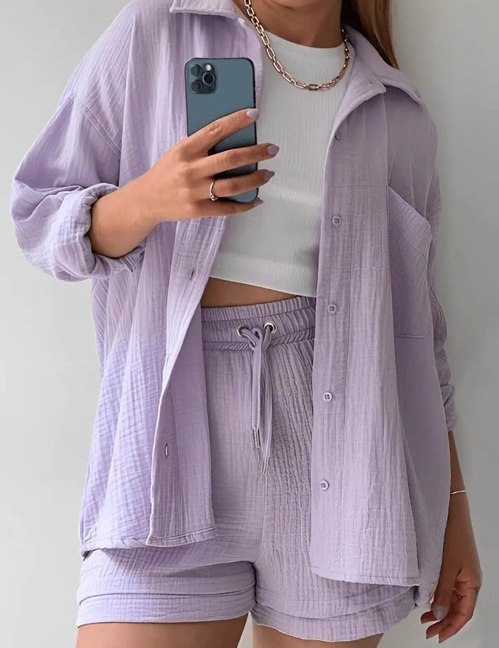 Donna con completo coordinato lilla, camicia e shorts in tessuto leggero, top bianco, collana dorata, smartphone in mano. Moda casual estiva.