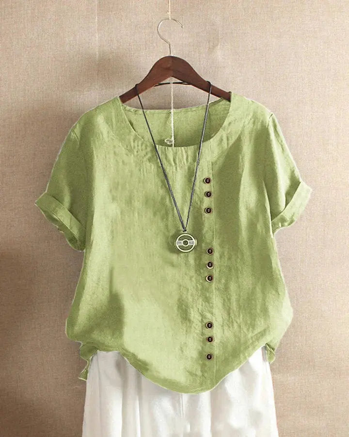 Camicia verde in lino con bottoni asimmetrici, maniche corte arrotolate, scollo rotondo, abbinata a pantaloni bianchi. Moda estiva casual.