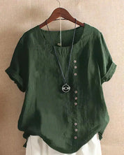 Blusa verde in lino con bottoni decorativi asimmetrici, maniche corte arrotolate, scollo rotondo, appesa su gruccia. Moda casual estiva donna.