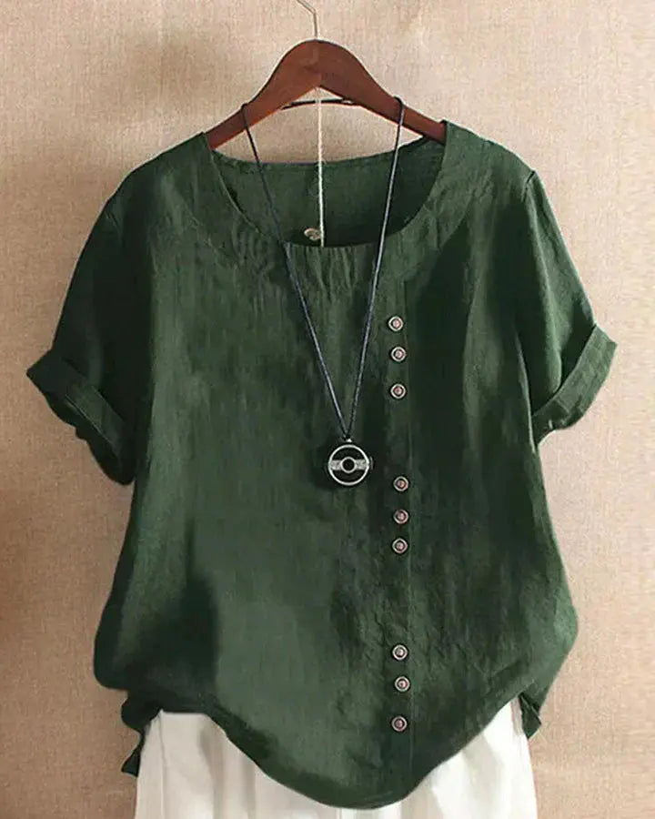 Blusa verde in lino con bottoni decorativi asimmetrici, maniche corte arrotolate, scollo rotondo, appesa su gruccia. Moda casual estiva donna.