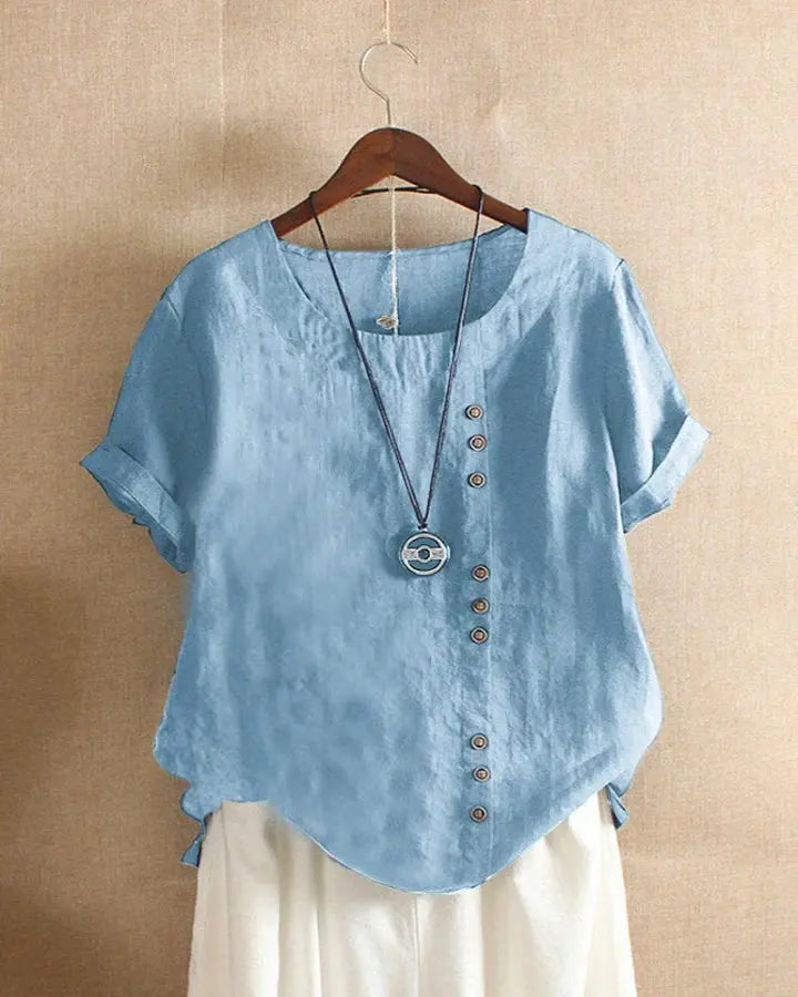 Camicia donna in lino azzurro con bottoni asimmetrici, maniche corte risvolto, abbinata a collana pendente, appesa su gruccia.