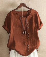 Maglietta marrone in lino con bottoni asimmetrici, maniche corte e collana decorativa, appesa su gruccia. Moda casual estiva donna.