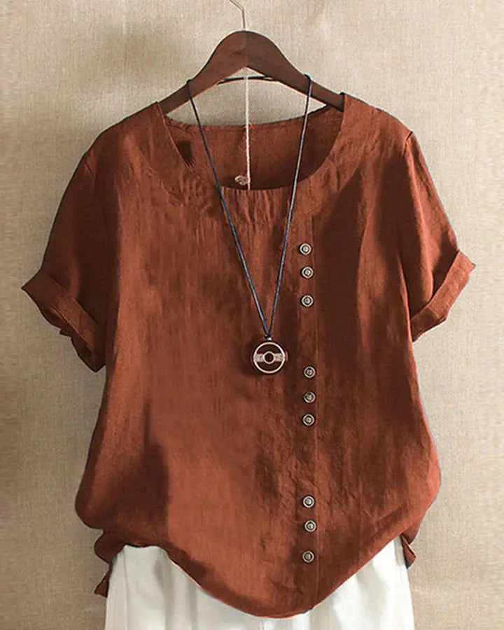 Maglietta marrone in lino con bottoni asimmetrici, maniche corte e collana decorativa, appesa su gruccia. Moda casual estiva donna.