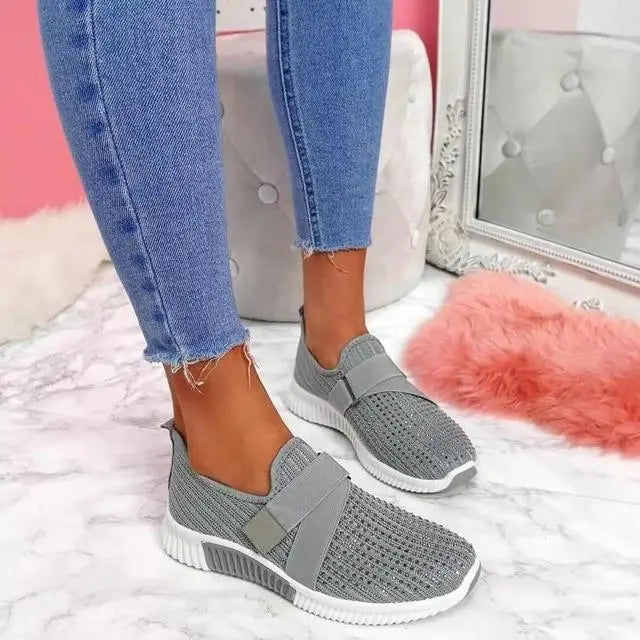 Scarpe da ginnastica grigie da donna su pavimento in marmo, indossate con jeans aderenti. Design traspirante, suola bianca, perfette per lo sport e il tempo libero.