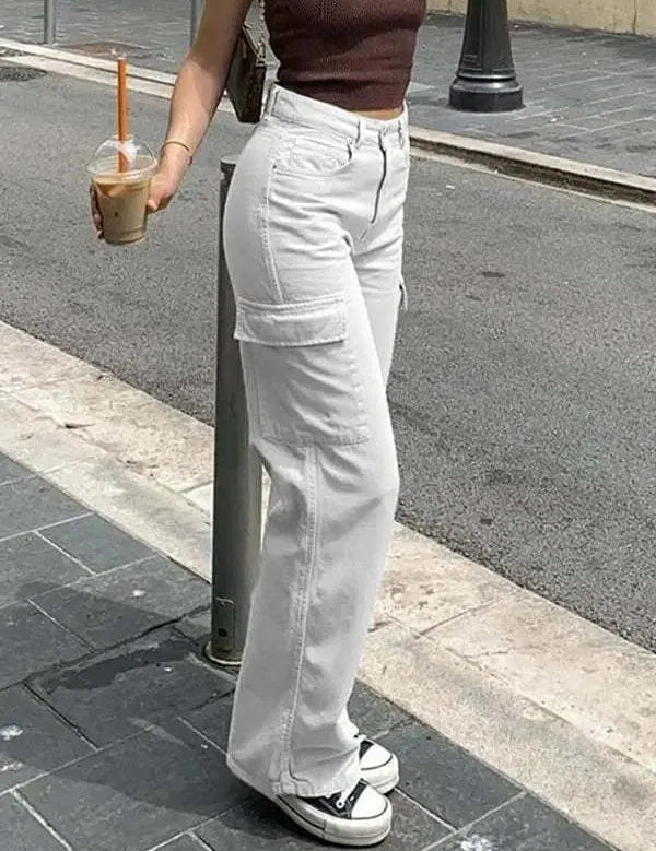 Donna con pantaloni cargo bianchi e top marrone, tiene un caffè freddo. Scarpe da ginnastica bianche e nere. Moda casual urbana.