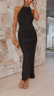 Abito lungo nero elegante senza maniche, donna in sandali beige, clutch marrone chiaro, sfondo muro in pietra, moda serale sofisticata.