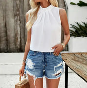 Donna con top bianco senza maniche e shorts di jeans strappati, accessori dorati, borsa di paglia. Moda estiva casual, look trendy e fresco.