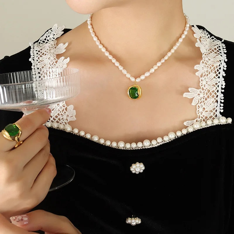 Collana di perle con ciondolo verde, anello coordinato, abito nero con dettagli in pizzo bianco, bicchiere di cristallo. Eleganza e stile classico.