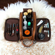 Beauty case in pelle marrone aperto su tappeto bianco, contenente pennelli trucco, flaconi cosmetici, rasoio e accessori per la cura personale.