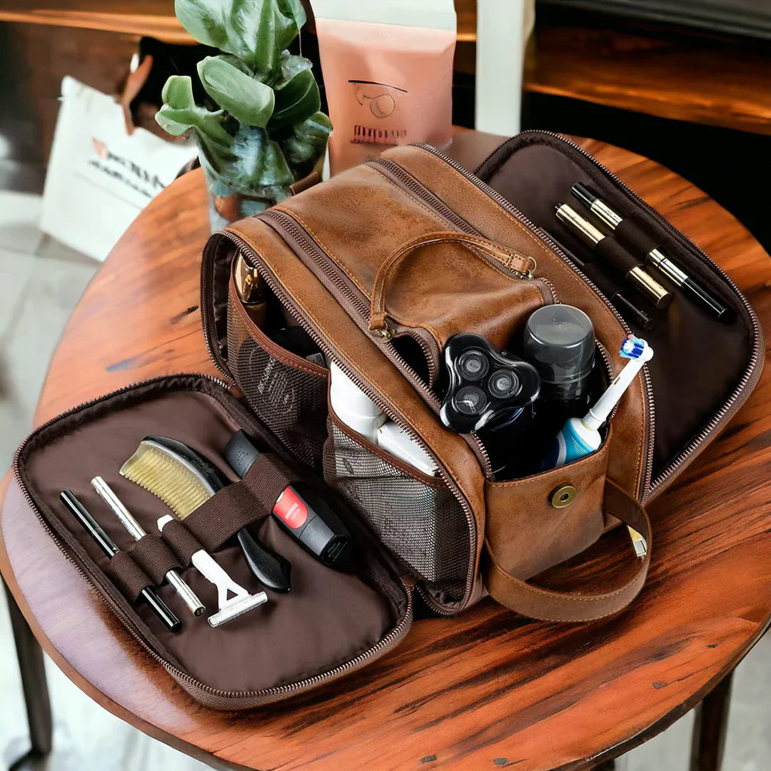 Borsa da toilette in pelle marrone aperta su tavolo, contenente articoli da toeletta come rasoio, spazzolino, pettine e flaconi. Accessori da viaggio uomo.