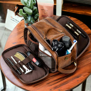 Borsa da toilette in pelle marrone aperta su tavolo, contenente articoli da toeletta come rasoio, spazzolino, pettine e flaconi. Accessori da viaggio uomo.