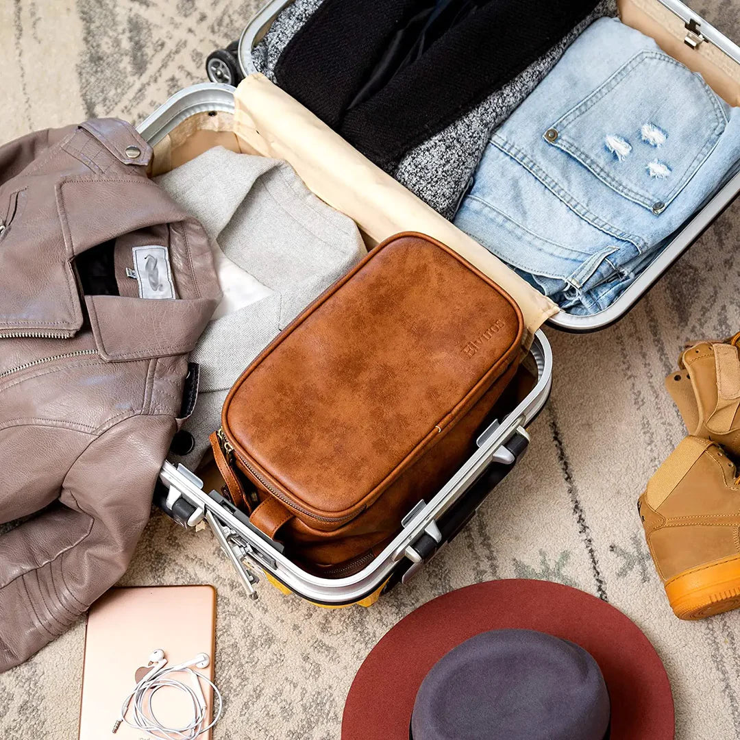 Valigia aperta con vestiti piegati, giacca in pelle, jeans, borsa marrone, cappello e scarpe su tappeto. Accessori da viaggio ordinati.
