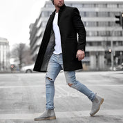 Uomo con cappotto nero elegante, jeans strappati azzurri e stivali grigi, cammina in città moderna. Moda urbana, abbigliamento invernale maschile.