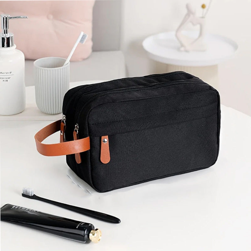 Borsa da toilette nera con manico in pelle marrone su tavolo bianco, accanto a spazzolino, dentifricio e dispenser sapone. Accessori bagno eleganti.