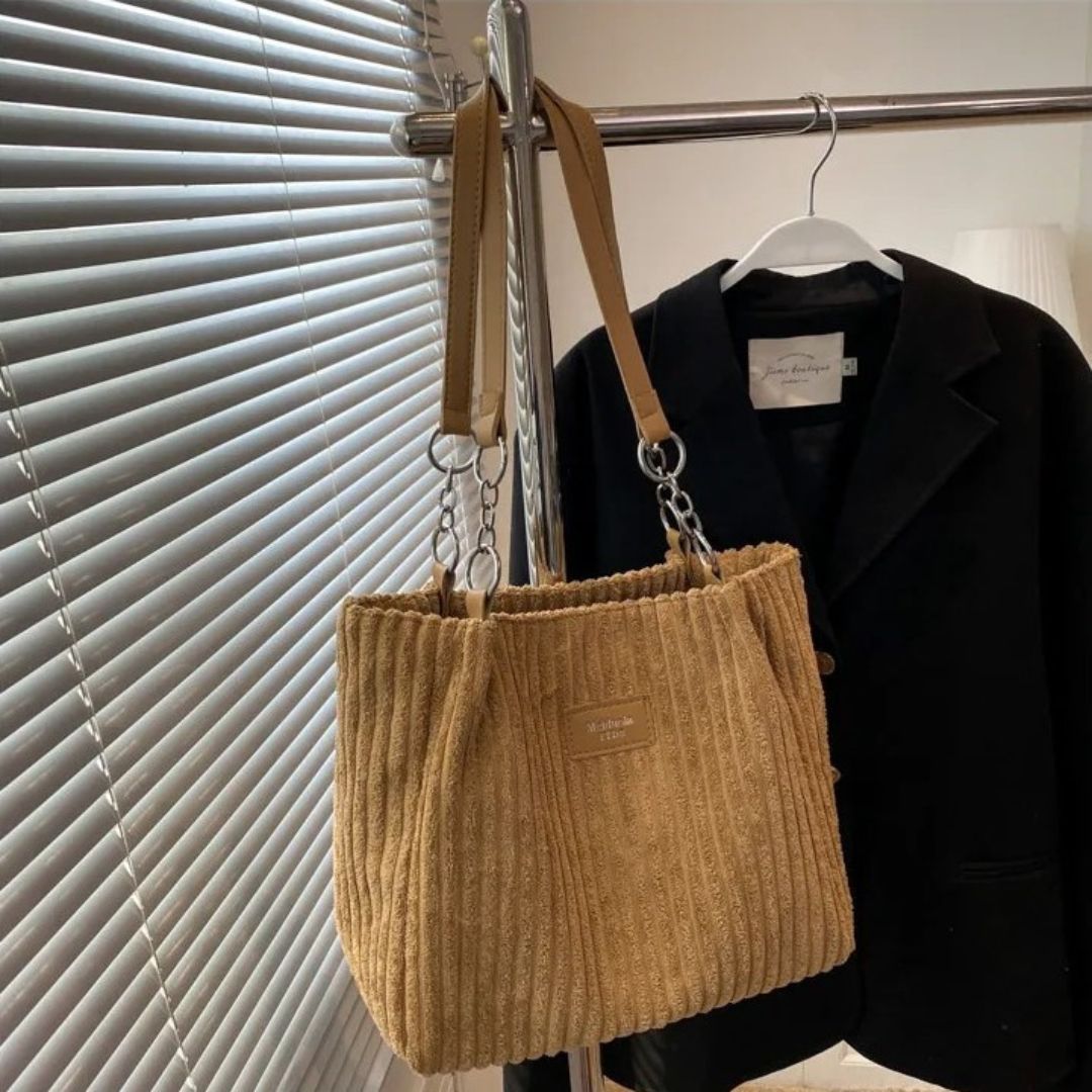 Borsa a spalla beige in tessuto a coste, appesa accanto a un blazer nero su un appendiabiti. Design elegante, perfetto per moda autunnale.