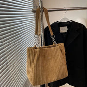 Borsa a spalla beige in tessuto a coste, appesa accanto a un blazer nero su un appendiabiti. Design elegante, perfetto per moda autunnale.