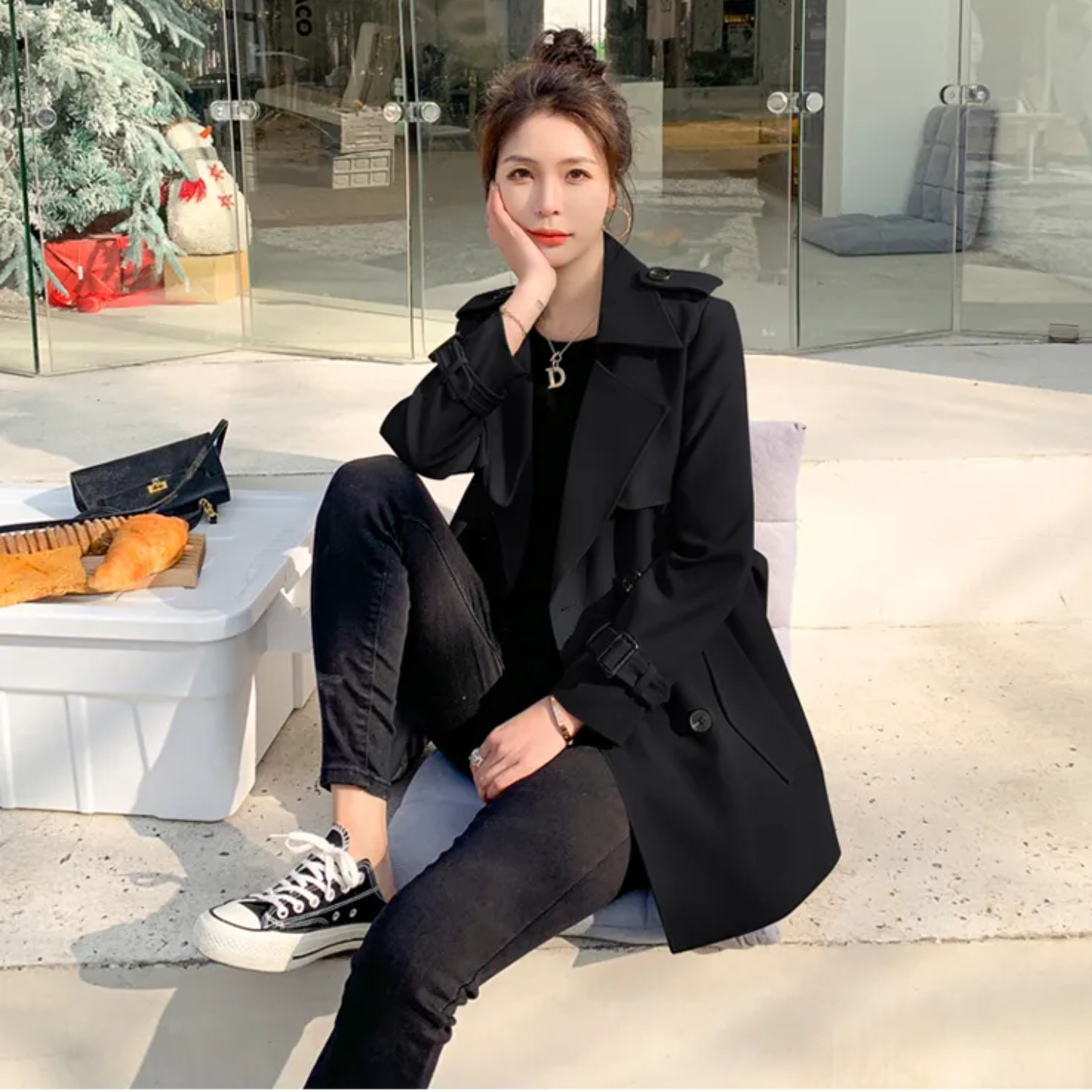 Donna seduta all'aperto con cappotto nero, jeans e sneakers Converse nere. Accanto a lei, una borsa e una scatola con baguette.