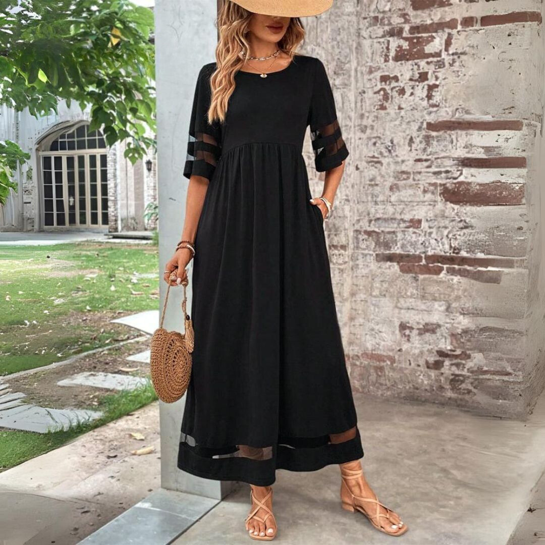 Donna con abito lungo nero elegante, maniche a tre quarti, borsa a tracolla rotonda, sandali beige, sfondo esterno con muro di mattoni.