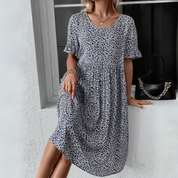 Donna con abito estivo floreale blu, maniche corte, accanto a borsa nera con catena dorata. Moda femminile casual elegante.