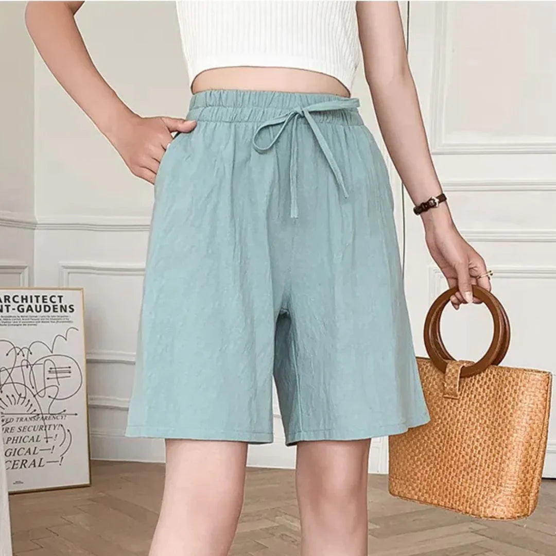 Donna con pantaloncini azzurri a vita alta e top bianco, tiene una borsa di paglia con manico circolare. Moda estiva casual elegante.