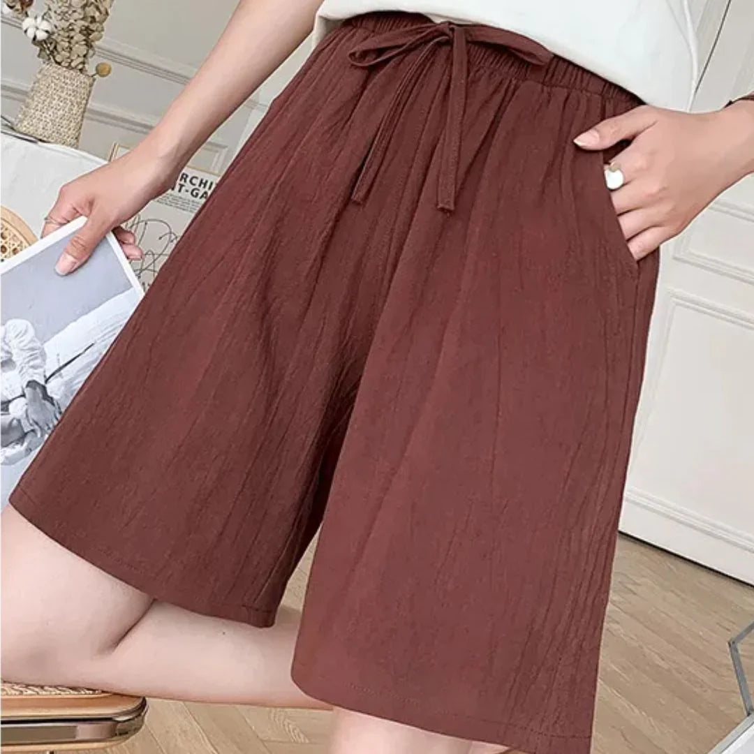 Shorts marroni da donna in tessuto leggero, con cintura regolabile, tasche laterali, indossati in un ambiente interno. Moda estiva casual.