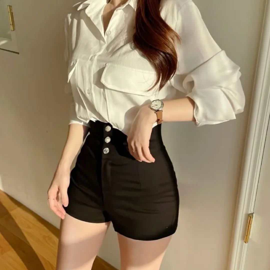 Donna in camicia bianca e pantaloncini neri a vita alta, orologio al polso, posa elegante. Moda femminile, abbigliamento casual chic.
