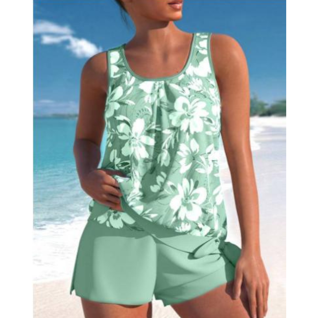 Donna in spiaggia con costume da bagno verde acqua a fiori bianchi, due pezzi, top senza maniche e pantaloncini. Mare e cielo blu sullo sfondo.