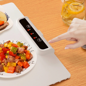 Piastra riscaldante elettrica bianca su tavolo in legno, con display digitale e piatto di verdure e carne. Mano che regola la temperatura.