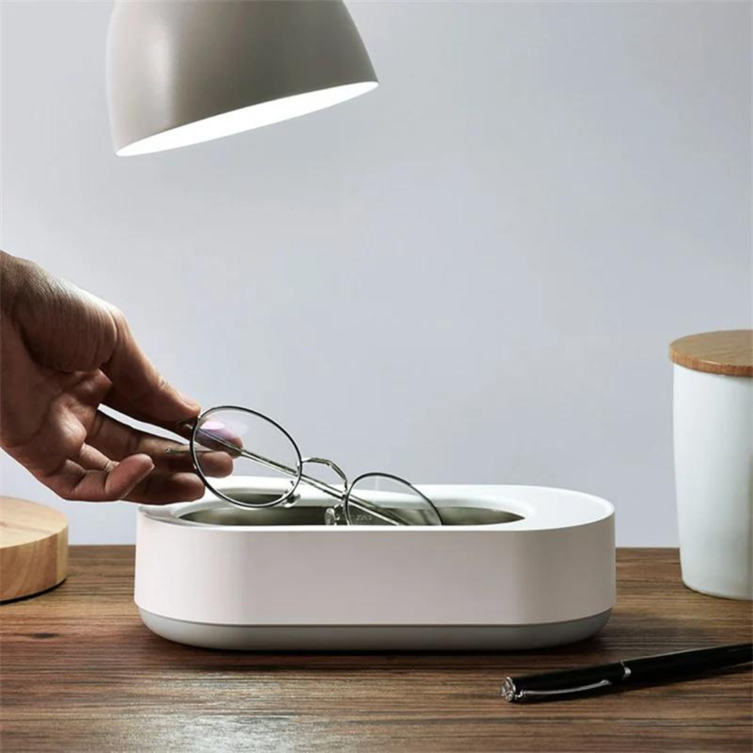 Occhiali in un pulitore ad ultrasuoni bianco su tavolo in legno, illuminato da lampada moderna. Accessorio per pulizia occhiali, design elegante.