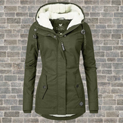 Giacca invernale verde oliva da donna con cappuccio foderato in pelliccia sintetica, chiusura a zip e bottoni, su sfondo di mattoni grigi.