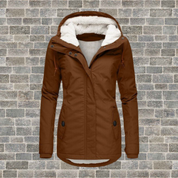 Giacca invernale da donna marrone con cappuccio foderato in pelliccia sintetica, chiusura a zip, tasche frontali, su sfondo di mattoni grigi.