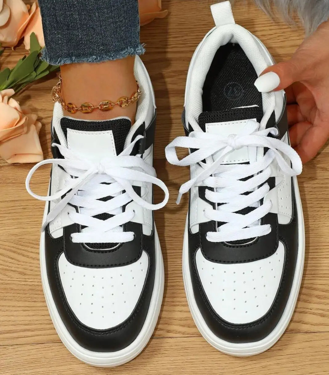 Scarpe da ginnastica bianche e nere su pavimento in legno, indossate con jeans e cavigliera dorata. Scarpe sportive moda donna, stile casual.