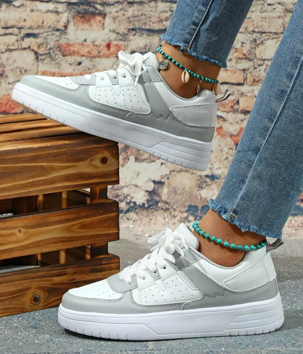 Scarpe da ginnastica bianche e grigie su sfondo di mattoni, indossate con jeans strappati e cavigliere turchesi. Moda casual, calzature trendy.