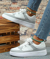 Scarpe da ginnastica bianche e grigie su sfondo di mattoni, indossate con jeans strappati e cavigliere turchesi. Moda casual, calzature trendy.