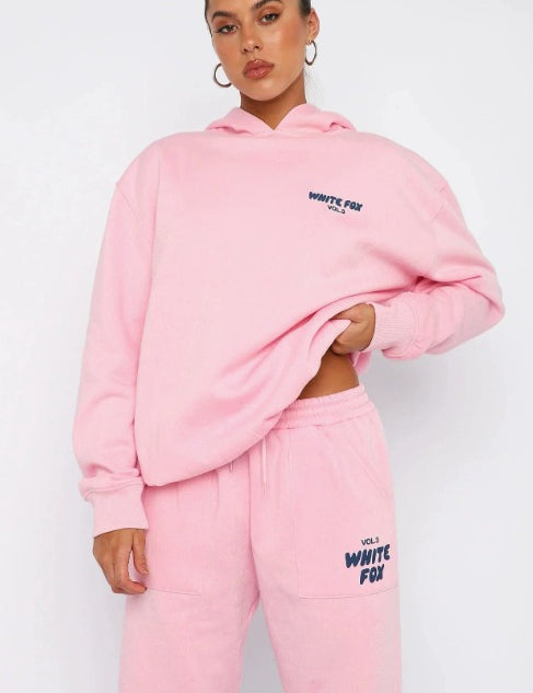 Donna con felpa e pantaloni rosa White Fox, logo visibile, posa casual. Abbigliamento sportivo alla moda, stile streetwear femminile.