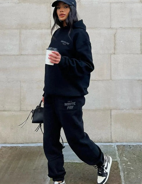 Donna con abbigliamento sportivo nero, felpa e pantaloni White Fox, cappellino da baseball, scarpe da ginnastica bianche e nere, tiene caffè e borsa.