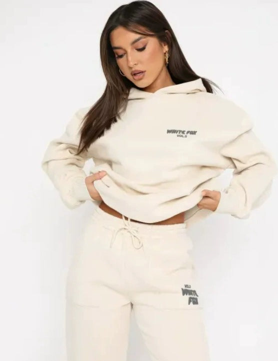 Modella indossa tuta sportiva beige con cappuccio, logo "White Fox" visibile. Abbigliamento casual donna, moda autunno, stile confortevole.