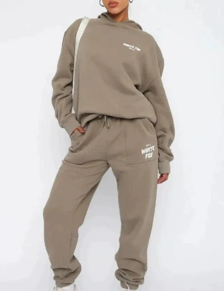 Donna in tuta sportiva beige, felpa con cappuccio e pantaloni abbinati, scarpe da ginnastica bianche. Moda casual, abbigliamento comodo.