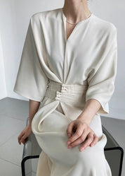 Abito elegante bianco avorio con cintura decorativa, maniche ampie e scollo a V, indossato da una donna seduta. Moda femminile sofisticata.