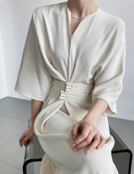 Abito elegante bianco avorio con cintura decorativa, maniche ampie e scollo a V, indossato da una donna seduta. Moda femminile sofisticata.