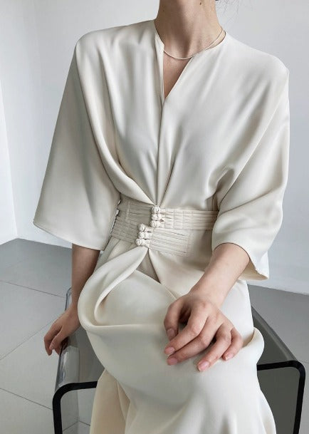 Abito elegante bianco avorio con cintura decorativa, maniche ampie e scollo a V, indossato da una donna seduta. Moda femminile sofisticata.
