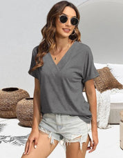 Donna con occhiali da sole indossa maglietta grigia a V e shorts di jeans strappati, seduta su divano esterno con cuscini e decorazioni in vimini.