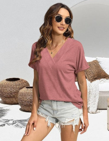 Donna con occhiali da sole indossa maglietta rosa a V e shorts di jeans strappati, sfondo con cesti di vimini e divano bianco. Moda estiva casual.