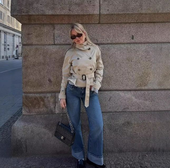 Donna con trench beige, jeans a zampa, occhiali da sole e borsa nera Chanel, appoggiata a un muro di pietra in città. Moda autunnale urbana.